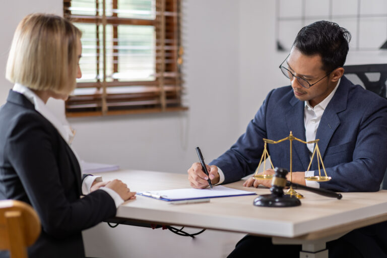 Lawyer signing documents at desk with client and scales of justice